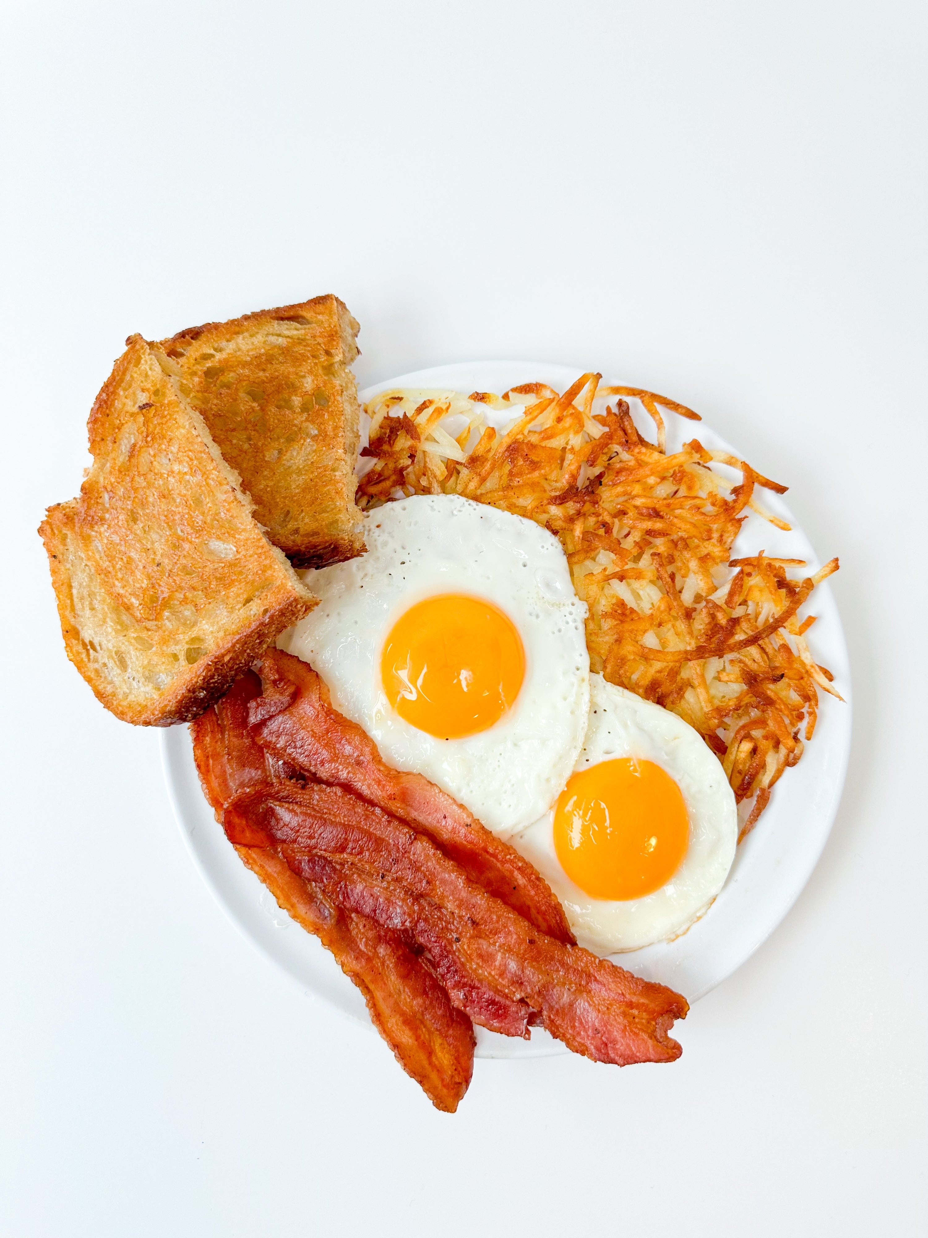 toast, bacon, sunny side up eggs, and hashbrowns on a white plate with a white background