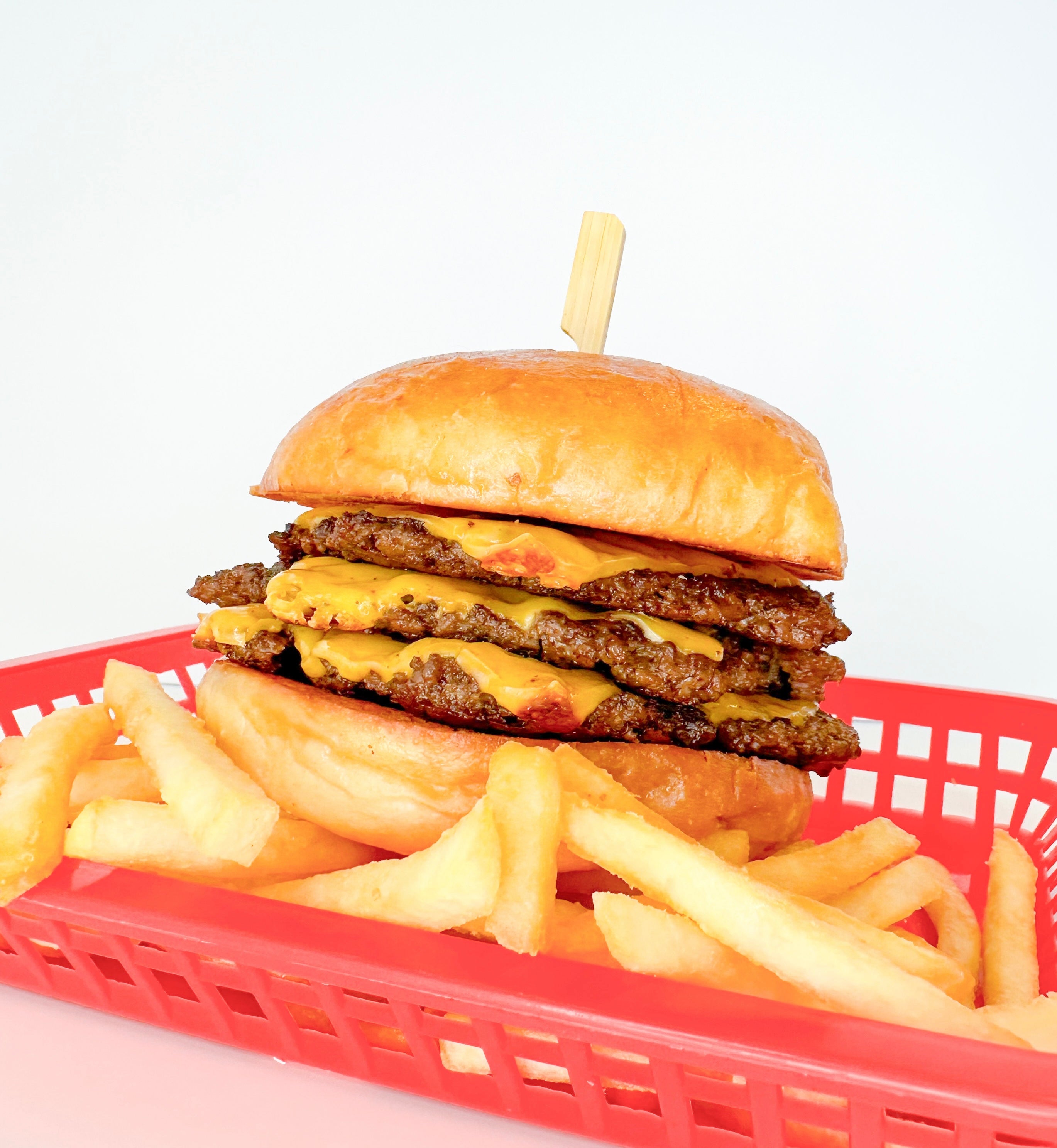 triple stack cheeseburger sitting on top of a bed of french fries in a red basket against a white background